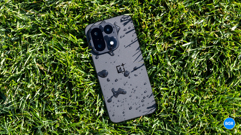 A OnePlus 15 smartphone resting in green grass with water droplets on its back panel.