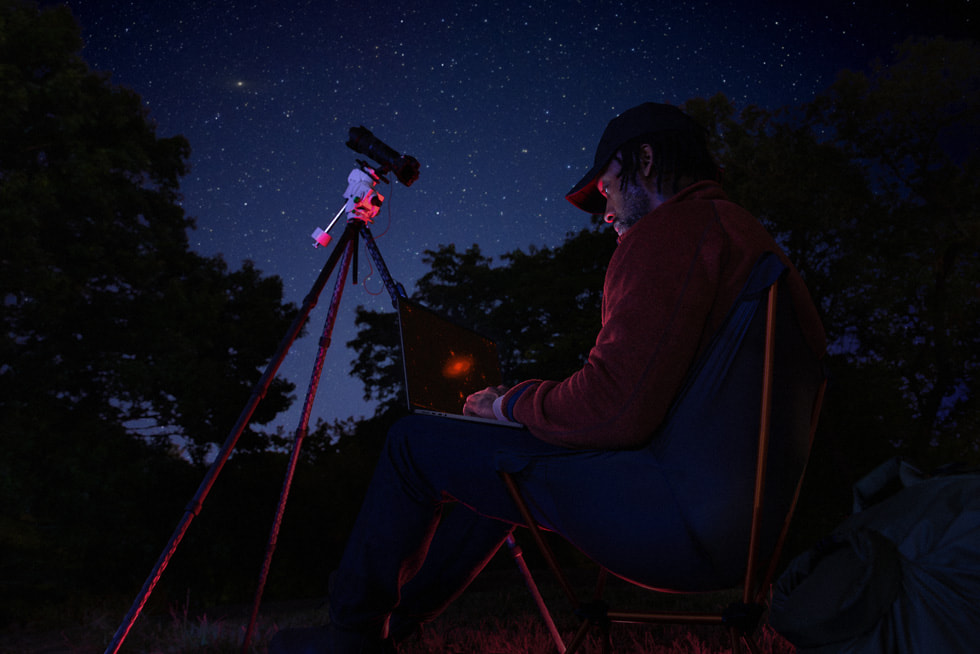 A user using the MacBook Pro outdoors at night alongside a telescope setup.