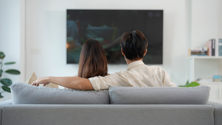A couple sitting comfortably on a couch in a dimly lit living room while watching television.