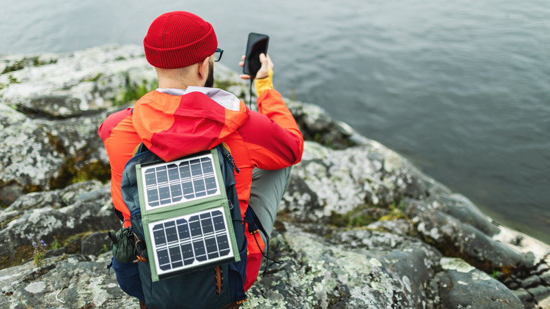 A hiker standing on a cliff with a solar power bank attached to their backpack, connected to a smartphone.