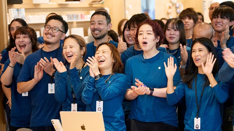 A group of Apple Store employees in blue shirts clapping and cheering at a store entrance.