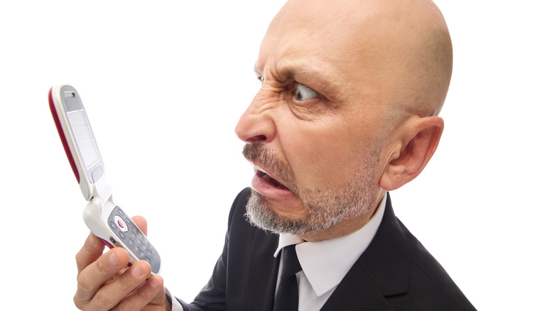 A middle-aged man looking frustrated while trying to read something on a small flip phone screen.