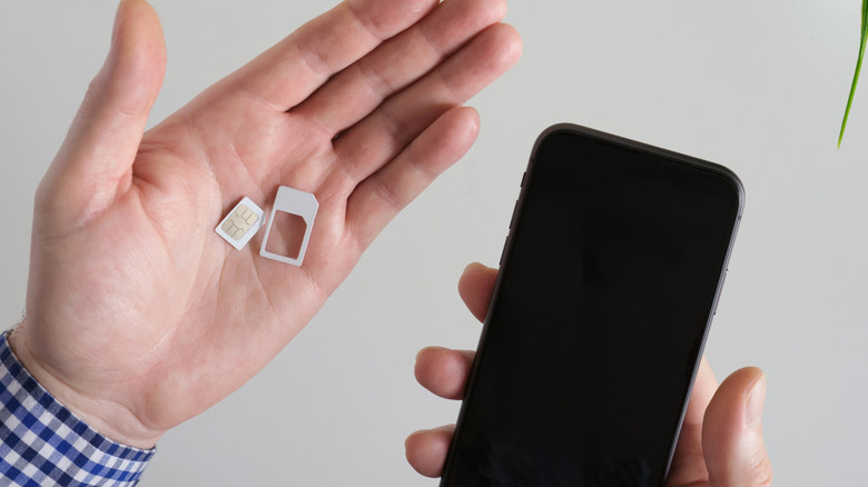 A hand holding a small physical SIM card next to a modern smartphone, symbolizing the hardware transition.